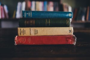 Four books piled up, red,yellow, black and blue spines in front of a book shelf. Blurb vs synopsis.