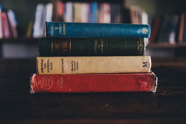 Four books piled up, red,yellow, black and blue spines in front of a book shelf. Blurb vs synopsis.